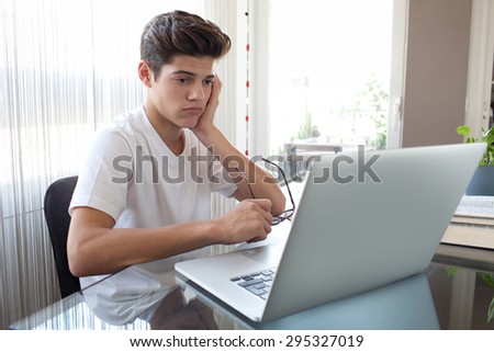 Teenager boy at a glass desk at home doing his homework using a laptop computer and books, holding reading glasses being unhappy, interior. Adolescent using technology, studying by window, indoors.