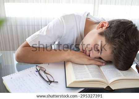 Teenager boy sleeping at a home  desk doing his homework using reading glasses and reference books, being tired, interior. Adolescent  preparing exams, studying by window with curtains, indoors.