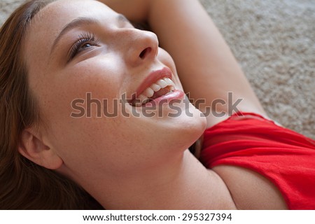 Close up beauty portrait of an attractive young woman laying down on a bed, wearing a bright red dress, looking happy and smiling joyfully, home interior. Beautiful woman lifestyle, relaxing indoors.