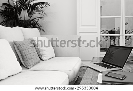 Still life black and white view of a living room with sofa, an open laptop computer with blank screen and a smartphone with paperwork, home interior. Aspirational technology indoors lifestyle.