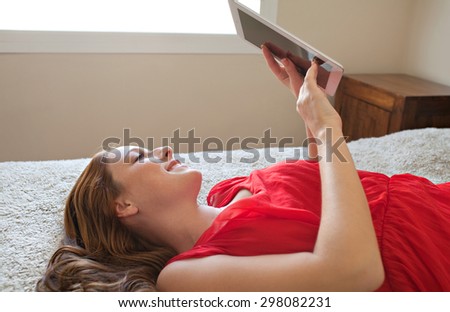 Side portrait of a beautiful young woman holding and using a digital tablet laying down on a bed at home, interior. Girl using technology, laying down relaxing and lounging in bedroom, indoors.