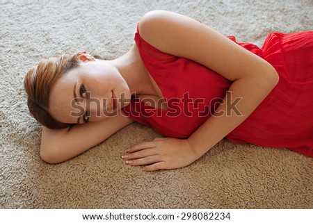 Beauty portrait of an attractive young woman laying on a bed at home wearing a bright red dress and gently smiling and looking at camera, home bedroom interior. Beauty feminine indoors lifestyle.