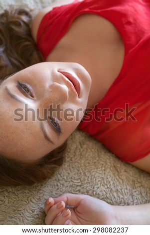 Beauty portrait of an attractive young woman laying on a bed at home wearing a bright red dress and gently smiling, looking away thoughtful, home bedroom interior. Beauty feminine indoors lifestyle.