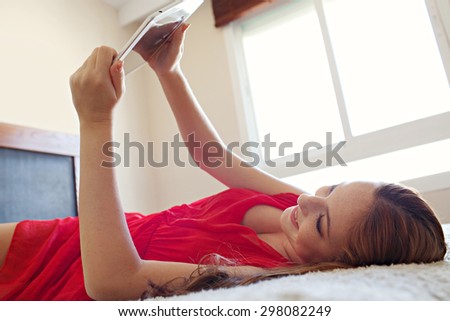 Side portrait of beautiful young woman holding and using a digital tablet laying down on a bed at home, interior. Girl using technology, laying down relaxing and lounging in bedroom, smiling indoors.