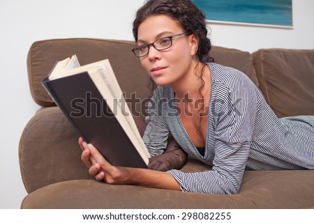 Portrait of beautiful young woman lounging on a sofa at home, reading a book wearing spectacles, living room interior. Relaxing home living lifestyle, indoors. Girl reading a book on the weekend.