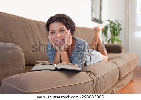 Beautiful and joyful young woman lounging on a comfortable sofa at home reading a book, laying down and relaxing in living room interior. Relaxing home lifestyle, indoors. Girl reading book, smiling.