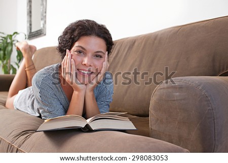 Beautiful and joyful young woman lounging on a comfortable sofa at home reading a book, laying down and relaxing in living room interior. Relaxing home lifestyle, indoors. Girl reading book, smiling.