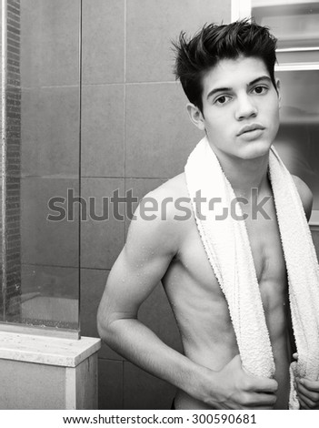 Black and white portrait of an adolescent young man drying after a shower at home, with white towel and thoughtfully looking at the camera, home bathroom interior. Healthy man lifestyle and grooming.