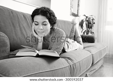 Black and white portrait of beautiful and serene young woman lounging on sofa at home reading a book, relaxing in living room interior. Relaxing home lifestyle, indoors. Girl reading book, smiling.