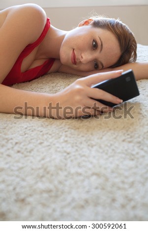 Portrait of attractive young woman laying on a bed at home, wearing a red dress, holding and using a smartphone while relaxing, home interior. Beautiful girl using technology in bedroom, interior.