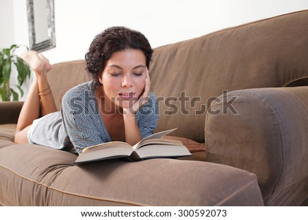 Beautiful and serene young woman lounging on a comfortable sofa at home reading a book, laying down and relaxing in living room interior. Relaxing home lifestyle, indoors. Girl reading book, smiling.