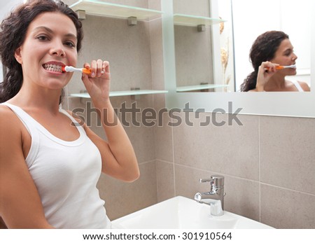 Beautiful young woman brushing her teeth in a home bathroom with her reflection on a mirror, home interior. Well being and healthy dental care lifestyle. Girl grooming indoors, morning routine.