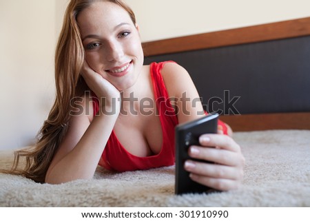 Portrait of attractive young woman laying on a bed smiling, wearing a red dress, using a smartphone relaxing, home interior. Beautiful girl using technology in bedroom looking at camera, interior.