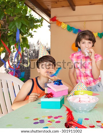 Two children brother and sister enjoying a birthday party at a celebratory fun table with food and sweets in a home garden, outdoors. Kids blowing party blowers, having fun together, home exterior.