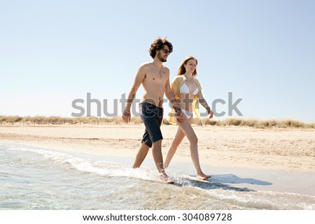 Honeymoon young couple holding hands relaxing and walking along the shore of sandy beach with rolling mountains and blue sea water and sky, outdoors. Travel tourism lifestyle, coastal destination.