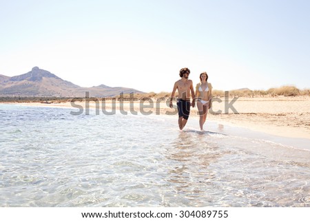 Honeymoon young couple holding hands relaxing and walking on the shore of a sandy beach with rolling mountains and blue sea water and sky, outdoors. Travel and tourism lifestyle, coastal destination.