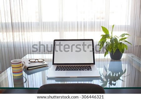 Still life view of an apartment living room with an open laptop on a professional glass desk workplace by a large bright window, interior. Home office technology, indoors. Working from home lifestyle.