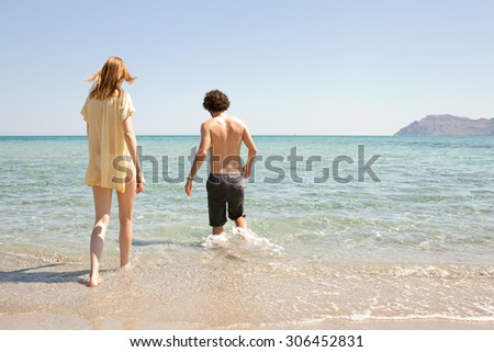 Rear view of young couple walking into the sea on holiday, on the shore of a white sand beach with blue sea water and sky, outdoors. Travel and tourism lifestyle, honeymoon coastal destination.