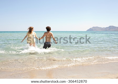 Rear view of young couple holding hands running into a clear sea on holiday, on the shore of a white sand beach with blue sky, outdoors. Travel and tourism lifestyle, honeymoon coastal destination.