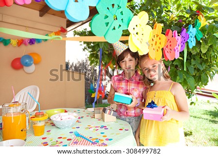Portrait of two young children girls friends together at a birthday party in a decorated home garden, smiling outdoors and holding gifts. Summer holiday celebration lifestyle.