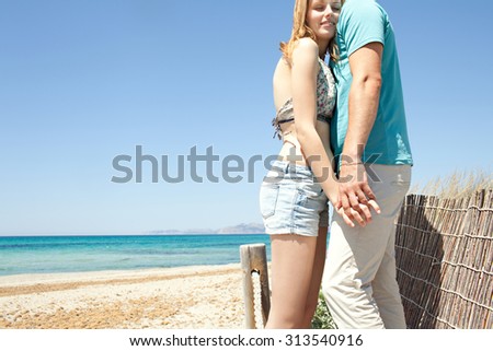 Attractive young tourist couple hugging with emotion on a sunny beach on a summer holiday destination, exterior. Boyfriend and girlfriend holding hands on vacation against a blue sky, outdoors.