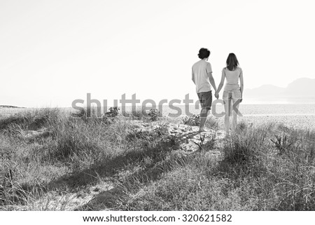 Black and white rear of a young tourist couple holding hands together in the distance on beach on holiday, contemplating outdoors. Travel and vacation lifestyle, nature living spacious exterior.
