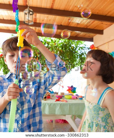 Kids having fun enjoying a colorful birthday party in a home garden, blowing bubbles and eating sweets, outdoors. Kid celebrating occasion in bright house exterior. Children fun activities, sunny day.