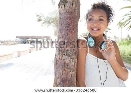 Portrait of a young African American black adolescent girl leaning on a tree trunk, smiling on beach holiday with headphones on a sunny day, outdoors. Teenager travel technology lifestyle, exterior.