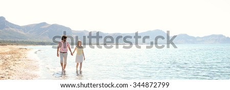 Panoramic view of tourist couple on holiday walking along the sea shore holding hands on vacation, sky with sun light, outdoors space. Romance and dynamic honeymoon calm lifestyle, summer exterior.