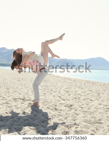Young tourist couple on holiday, with man carrying girl on back with legs up, on beach destination with sunny blue sky, outdoors space. Romance and dynamic honeymoon fun lifestyle, summer exterior.