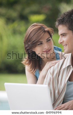 Couple looking at each other while using a laptop computer in home garden by swimming pool.