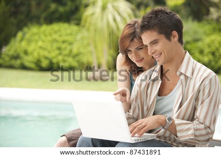 Couple using a laptop by swimming pool, hotel exterior.