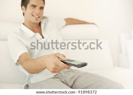 Young man using a tv control remote while sitting on a white leather sofa at home.