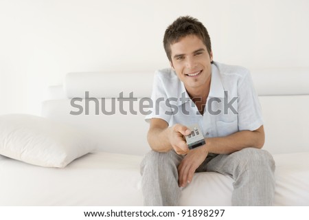 Young man using a tv control remote while sitting on a white leather sofa at home.