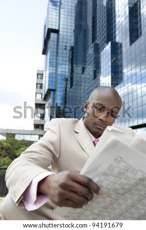 Businessman reading the newspaper while sitting down by a modern glass building in the city.