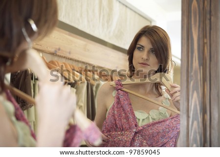 Sophisticated woman looking at herself in the mirror of a store, while holding a dress in front of her.