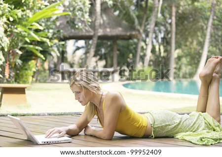 Attractive blonde woman using a laptop computer while laying down on a wooden deck in a tropical garden.