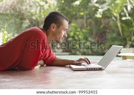 Young man using a laptop computer while laying down in a garden with a swimming pool.