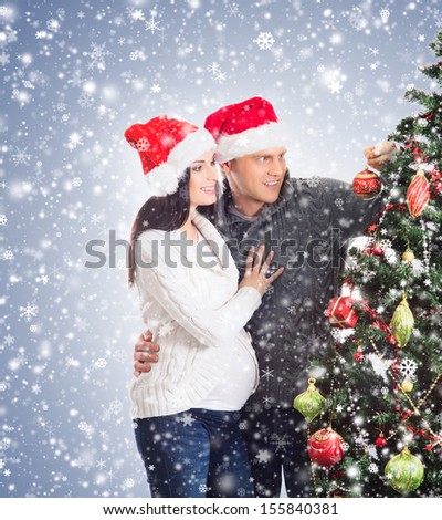 Young pregnant woman and happy father decorating Christmas tree over blue background with snow