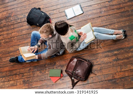 Girl and boy reading books leaning on each other on wooden floor having notebooks and bags around them.