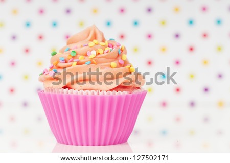Pink cupcake with whipped cream and sprinkles on a rainbow dotted background