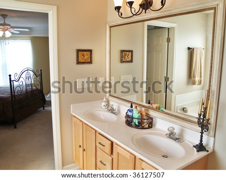 A modern luxurious bathroom with a large in an American home. A bed is visible through the doorway in the background.