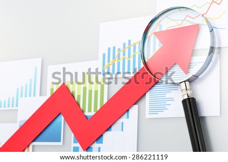 Red arrow and magnifying glass on the background of graphs and charts. Looking upside growth arrow with magnifying glass. Graphs and data confirming growth.