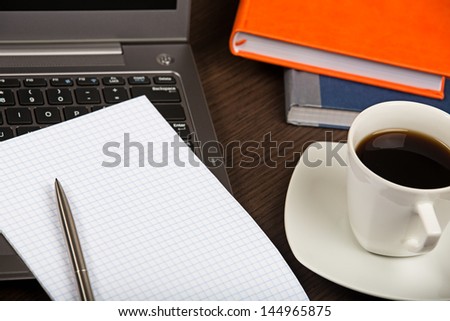 Laptop, sheet of paper, books, pen and cup of coffee on dark wooden office desk.