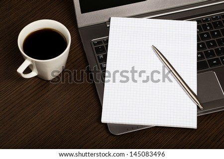 Top view on dark wooden office desk with laptop, sheet of paper, pen and cup of coffee.