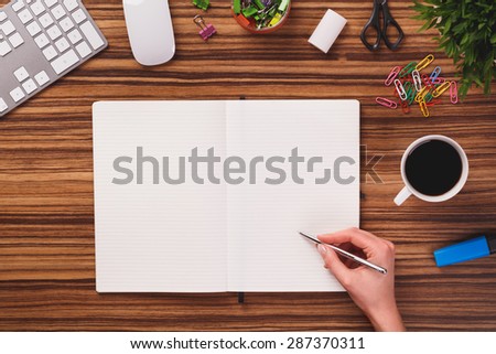 Hand holding a pen and writing something in opened notebook, which is lying next to computer keyboard, mouse, mug of black coffee and other office equipment on dark wooden office desk.
