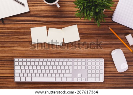 Modern white keyboard, mouse, sticky notes, notepad, pencil, mug of coffee, plant and other office equipment on dark wooden desk.