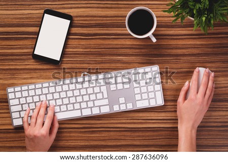 Someone is using computer keyboard and mouse next to modern black smartphone with empty screen, cup of black coffee and plant od dark wooden office desk.