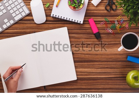 Someone is writing on notebook with pen on dark wooden office desk with modern computer keyboard, mouse notepad, highlighters, cup of coffee, apple and other.
