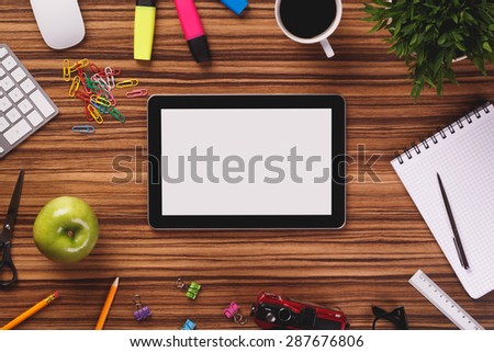 Modern black digital tablet with blank screen in the middle of office equipment such as computer keyboard, mouse, cup of coffee, notepad, apple, pencil, highlighters on dark wooden office desk.